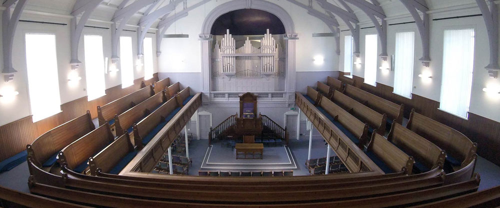 Interior of a church with wooden pews, pulpit, and pipe organ, illuminated by wall sconces